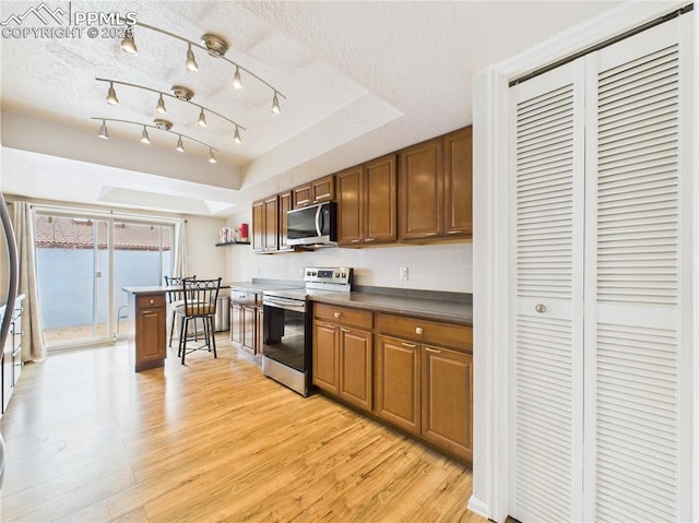 kitchen with a textured ceiling, brown cabinets, appliances with stainless steel finishes, light wood-type flooring, and a tray ceiling