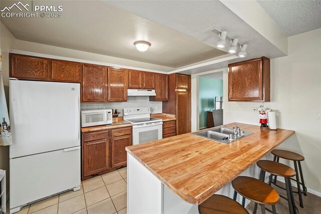 kitchen featuring white appliances, light tile patterned floors, under cabinet range hood, a sink, and a peninsula