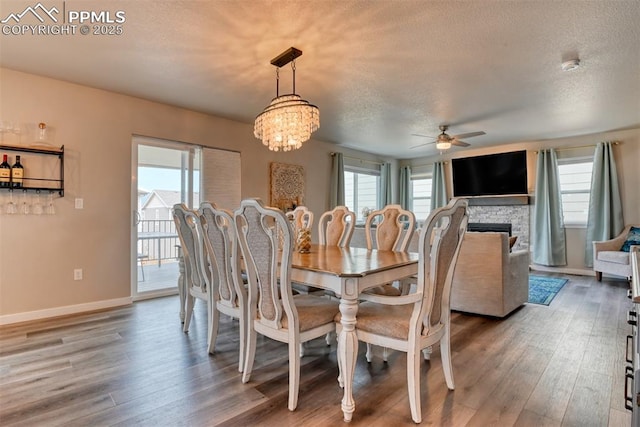dining area featuring ceiling fan with notable chandelier, a textured ceiling, a stone fireplace, wood finished floors, and baseboards