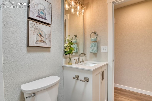 half bath with vanity, toilet, wood finished floors, a textured wall, and baseboards