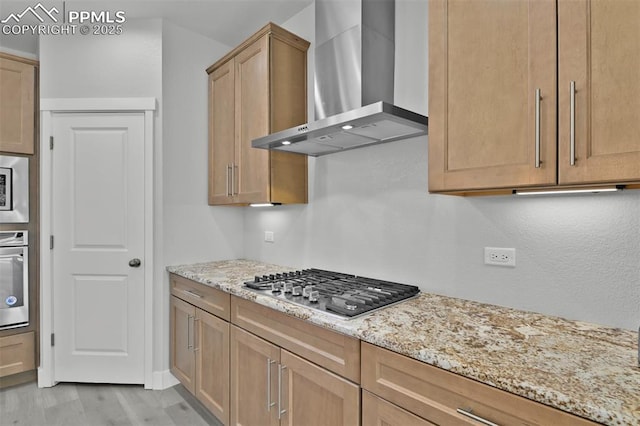 kitchen featuring wall chimney range hood, stainless steel appliances, light wood-style flooring, and light stone counters