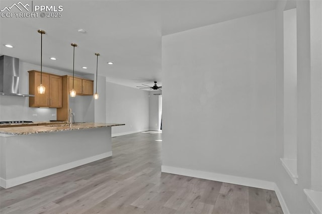 kitchen with light wood-style floors, ceiling fan, light stone countertops, wall chimney range hood, and brown cabinets