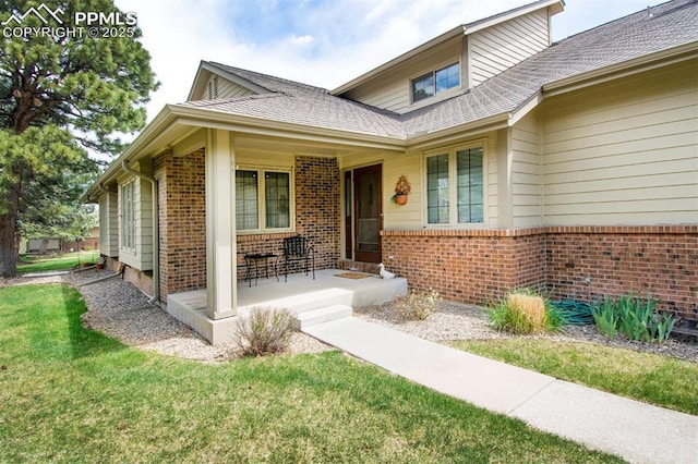 doorway to property featuring roof with shingles, covered porch, a yard, and brick siding