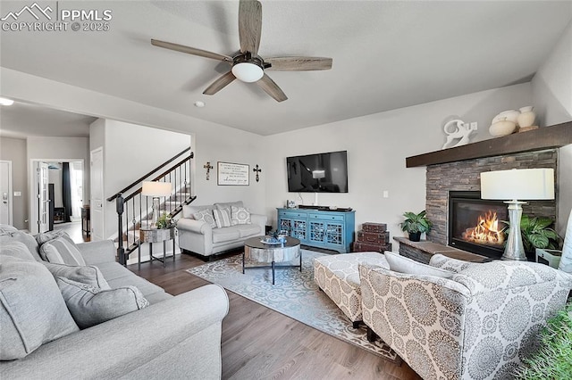 living room with a stone fireplace, a ceiling fan, wood finished floors, and stairs