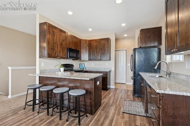 kitchen with a sink, light wood-type flooring, a kitchen bar, light stone counters, and black appliances