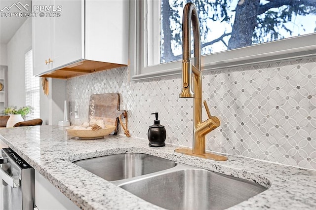 interior details featuring stainless steel dishwasher, a sink, backsplash, white cabinetry, and light stone counters