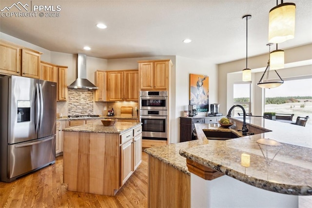 kitchen featuring light wood finished floors, a sink, wall chimney exhaust hood, a center island, and appliances with stainless steel finishes