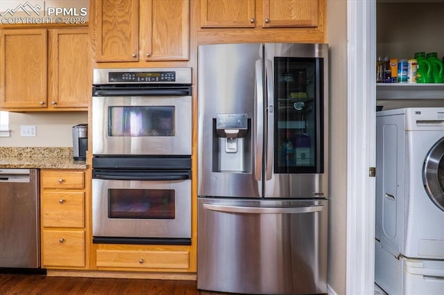 kitchen featuring brown cabinetry, appliances with stainless steel finishes, washing machine and dryer, dark wood-style flooring, and light stone countertops