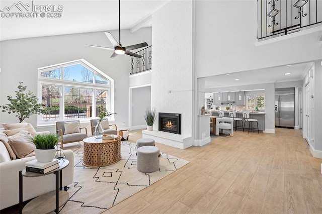 living area with high vaulted ceiling, a ceiling fan, light wood-style floors, baseboards, and a brick fireplace