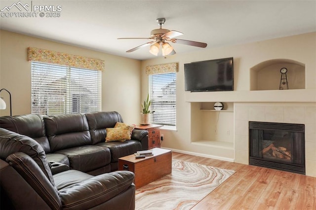 living area with ceiling fan, a tiled fireplace, built in shelves, wood finished floors, and baseboards