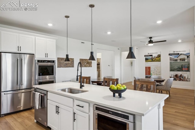kitchen with light wood-type flooring, an island with sink, ceiling fan, stainless steel appliances, and a sink