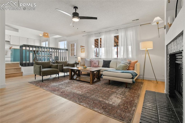 living area featuring a textured ceiling, light wood-style floors, a brick fireplace, a ceiling fan, and stairs