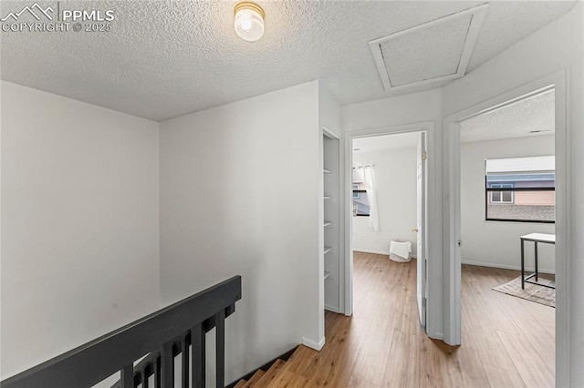 hall featuring attic access, baseboards, light wood-style floors, and a textured ceiling