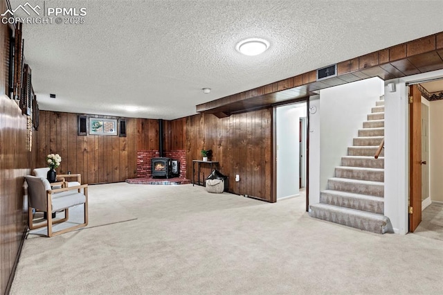 sitting room with carpet floors, a wood stove, stairs, wooden walls, and a textured ceiling