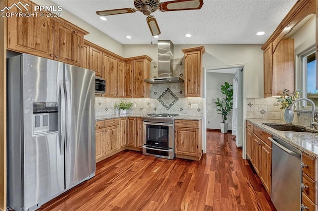 kitchen featuring wall chimney range hood, a sink, ceiling fan, backsplash, and appliances with stainless steel finishes