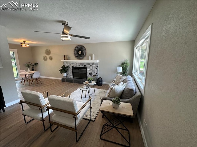 living room with ceiling fan, a tiled fireplace, wood finished floors, baseboards, and a textured wall