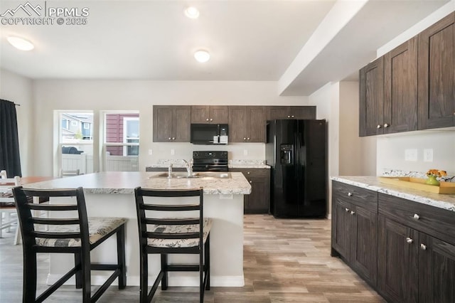kitchen with dark brown cabinetry, a breakfast bar area, a kitchen island with sink, light wood-type flooring, and black appliances