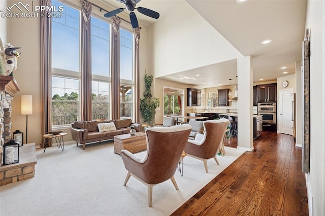 living area featuring ceiling fan, dark wood-style floors, recessed lighting, a stone fireplace, and a high ceiling