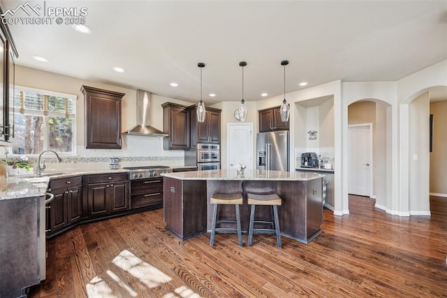 kitchen featuring tasteful backsplash, appliances with stainless steel finishes, a center island, dark brown cabinetry, and wall chimney exhaust hood