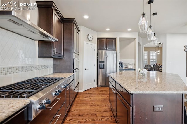 kitchen featuring dark brown cabinetry, wood finished floors, decorative backsplash, appliances with stainless steel finishes, and wall chimney exhaust hood