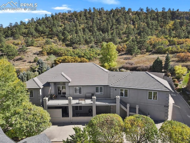 back of house with a tile roof, a view of trees, a balcony, and stucco siding