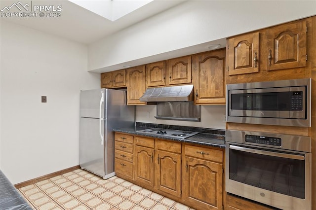kitchen featuring brown cabinetry, dark countertops, light floors, under cabinet range hood, and appliances with stainless steel finishes