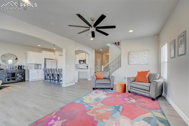 living room with ceiling fan, visible vents, arched walkways, light wood finished floors, and stairs