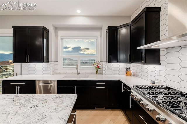 kitchen with light stone countertops, dark cabinets, appliances with stainless steel finishes, a sink, and wall chimney exhaust hood