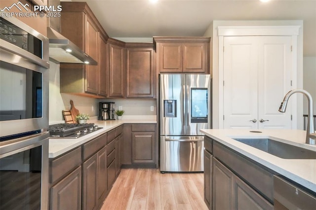 kitchen featuring appliances with stainless steel finishes, light wood-style flooring, a sink, light countertops, and wall chimney exhaust hood