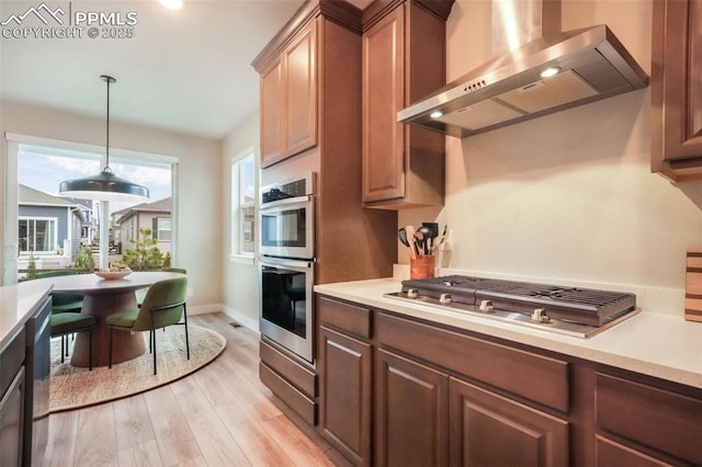 kitchen with appliances with stainless steel finishes, light wood-style floors, light countertops, wall chimney exhaust hood, and decorative light fixtures