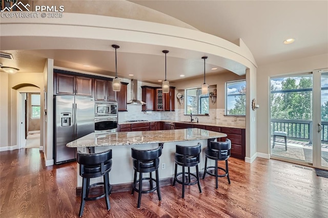 kitchen featuring wall chimney range hood, appliances with stainless steel finishes, light stone counters, backsplash, and glass insert cabinets