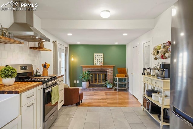 kitchen with open shelves, wall chimney exhaust hood, wood counters, appliances with stainless steel finishes, and light tile patterned floors