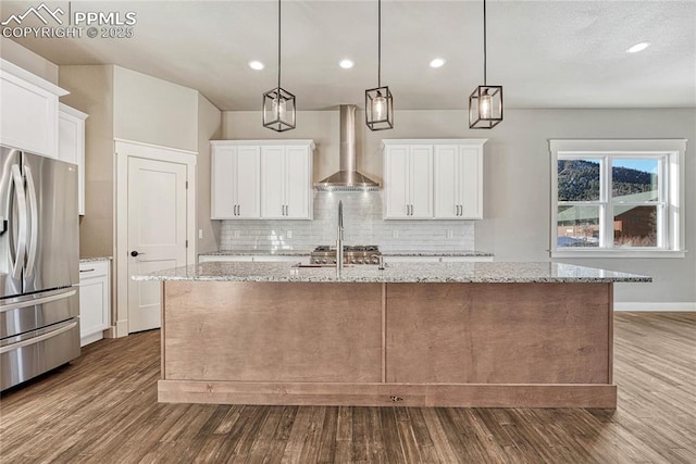 kitchen with stainless steel refrigerator with ice dispenser, a center island with sink, dark wood finished floors, wall chimney range hood, and tasteful backsplash