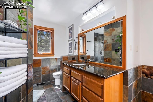 bathroom featuring stone tile flooring, vanity, toilet, a wainscoted wall, and a tile shower