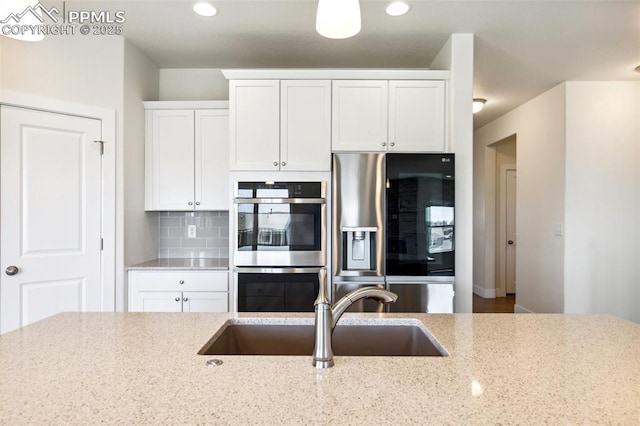 kitchen featuring refrigerator with ice dispenser, a sink, stainless steel double oven, white cabinets, and light stone countertops