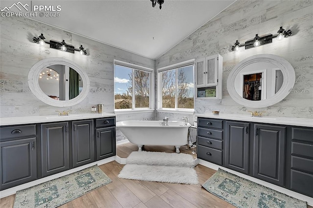 bathroom featuring a freestanding bath, vaulted ceiling, wood finished floors, two vanities, and wallpapered walls