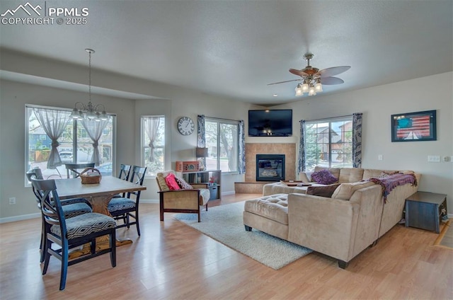 living area featuring baseboards, a tiled fireplace, light wood-style flooring, and ceiling fan with notable chandelier
