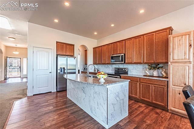 kitchen featuring light stone counters, a sink, arched walkways, brown cabinets, and appliances with stainless steel finishes
