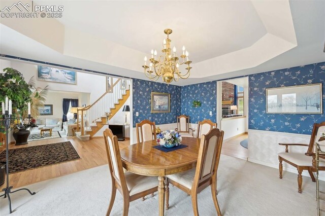 carpeted dining area with wallpapered walls, stairs, a wainscoted wall, a chandelier, and a raised ceiling