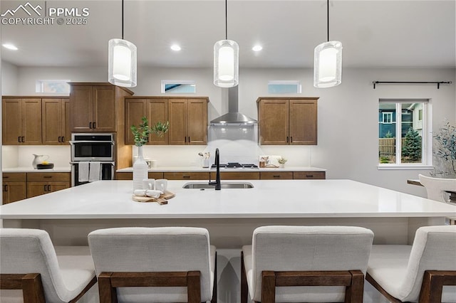 kitchen featuring wall chimney exhaust hood, a sink, stainless steel double oven, brown cabinetry, and recessed lighting