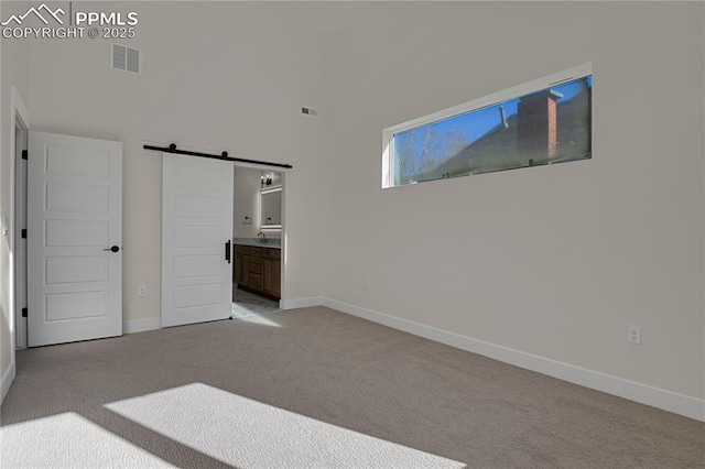 unfurnished bedroom with a barn door, light carpet, baseboards, a high ceiling, and ensuite bathroom