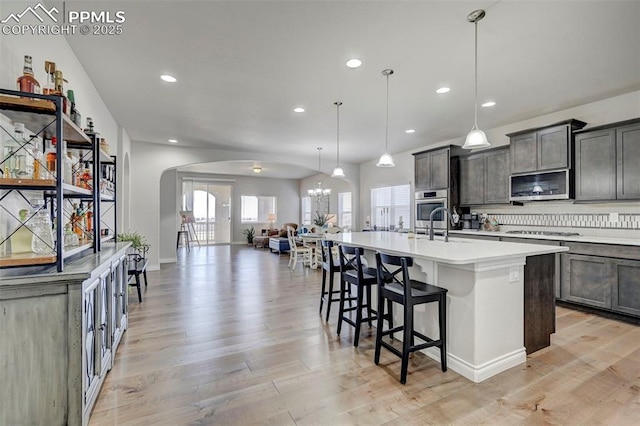 kitchen with appliances with stainless steel finishes, arched walkways, open floor plan, a center island with sink, and light wood-style flooring