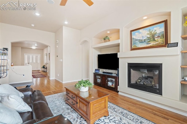 living area with arched walkways, light wood-style flooring, built in features, a ceiling fan, and a glass covered fireplace