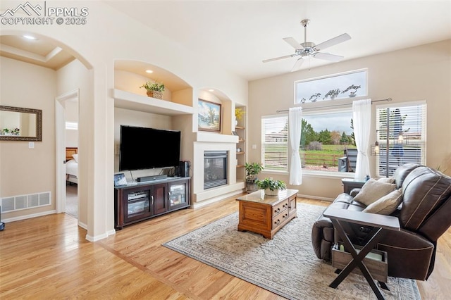 living area featuring light wood-type flooring, built in shelves, a ceiling fan, a glass covered fireplace, and baseboards