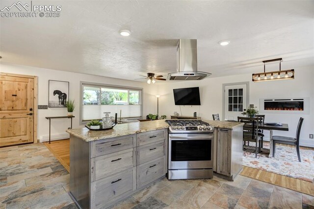 kitchen with stainless steel gas range, wall chimney range hood, gray cabinetry, a ceiling fan, and light stone finish flooring
