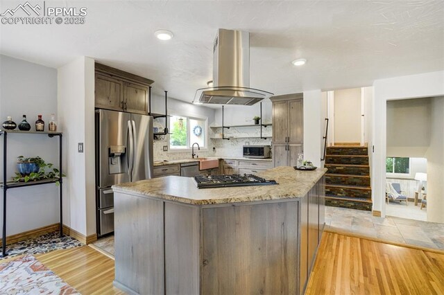 kitchen featuring stainless steel appliances, island exhaust hood, decorative backsplash, light wood-style flooring, and baseboards