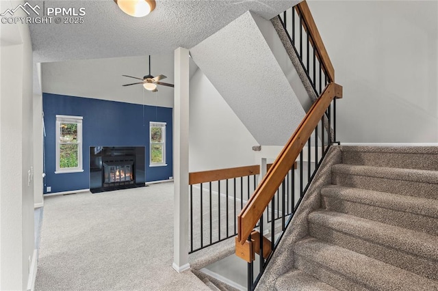 stairs featuring a ceiling fan, lofted ceiling, a textured ceiling, carpet floors, and a fireplace with flush hearth