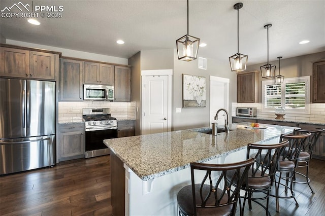 kitchen with appliances with stainless steel finishes, a sink, backsplash, dark wood finished floors, and light stone countertops