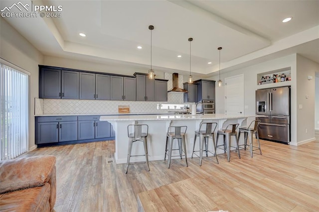 kitchen featuring high end refrigerator, a tray ceiling, wall chimney exhaust hood, light countertops, and a kitchen breakfast bar