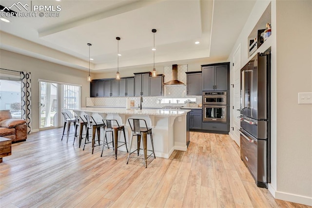 kitchen featuring appliances with stainless steel finishes, wall chimney range hood, a tray ceiling, decorative backsplash, and light wood-style flooring
